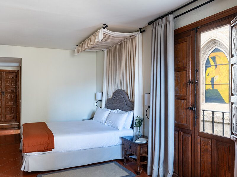 Habitación acogedora con cama blanca, dosel beige, mesita de noche y vidriera visible a través de puertas de madera.