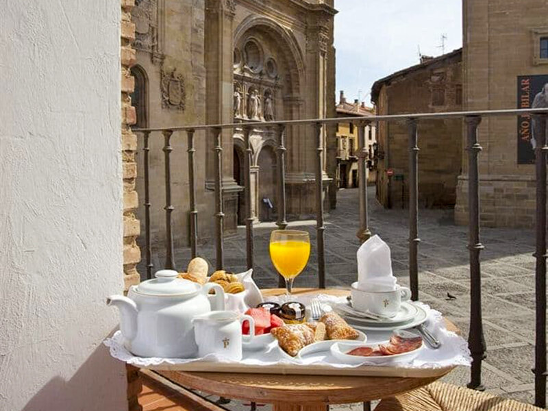 Una bandeja de desayuno con zumo, café y bollería en un balcón con vistas a una calle histórica.