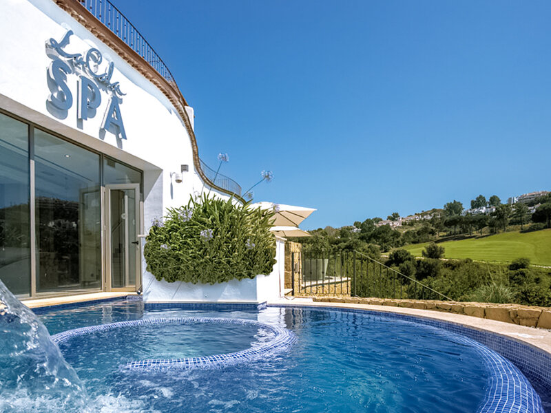 Un jacuzzi al aire libre frente a un edificio blanco de spa con una colina verde al fondo.