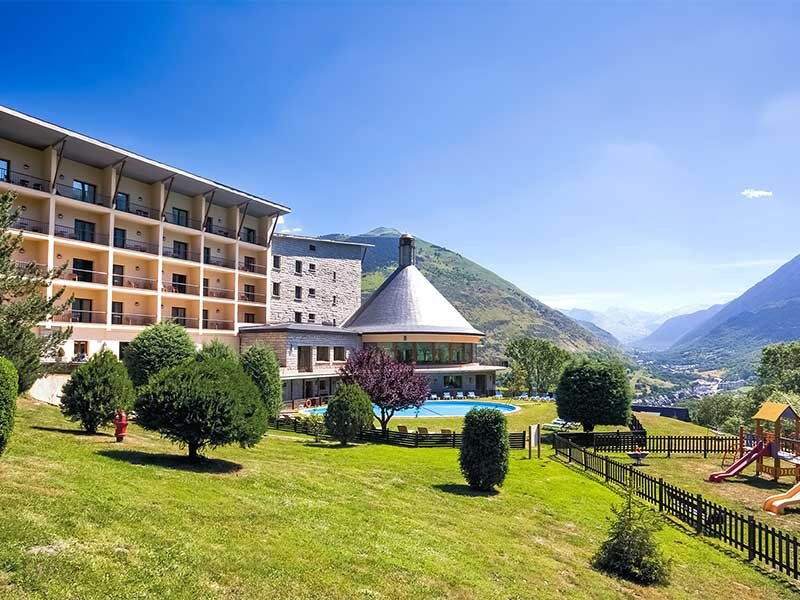 Un gran hotel con piscina, césped y parque infantil frente a un paisaje montañoso bajo el cielo azul.