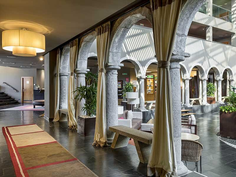 Un pasillo interior muestra arcos de piedra, cortinas, plantas en macetas y zonas de estar modernas.
