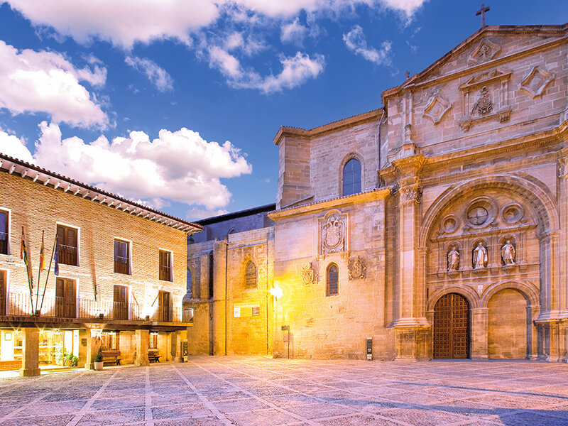 Plaza iluminada ante una iglesia histórica y un edificio antiguo bajo un cielo azul con nubes.