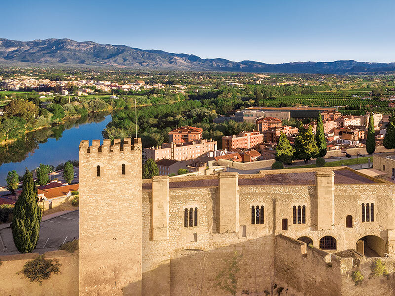 Antigua fortaleza de piedra con torre, río y ciudad al fondo bajo un cielo azul.