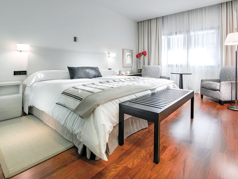 Dormitorio moderno y luminoso con cama grande, suelo de madera oscura, banco negro y sillones grises.