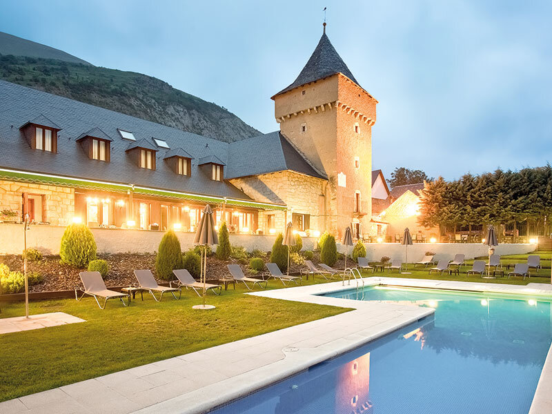 Un resort de piedra iluminado con torre, piscina y tumbonas en primer plano al atardecer.