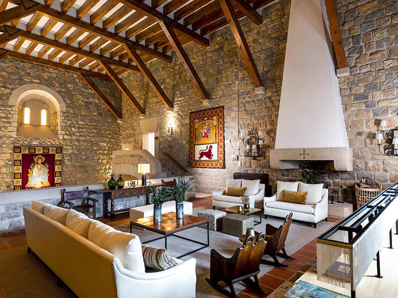 Acogedora sala con paredes de piedra, techo de vigas de madera, gran chimenea, sofás blancos y tapices medievales.