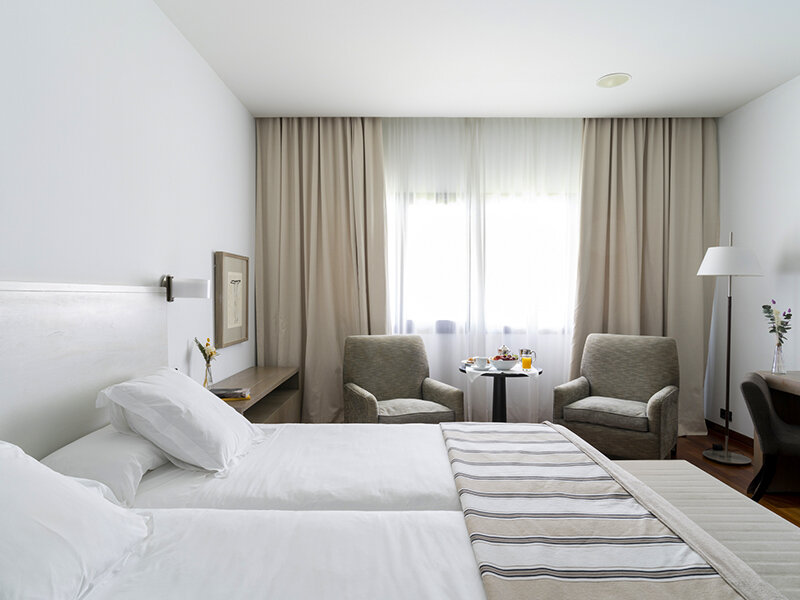 Una habitación de hotel luminosa con dos camas, sillones y una mesita con desayuno junto a la ventana.