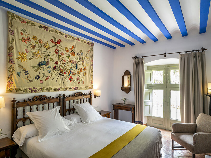 Un dormitorio con cama blanca, camino amarillo, gran tapiz floral y vigas azules en el techo.