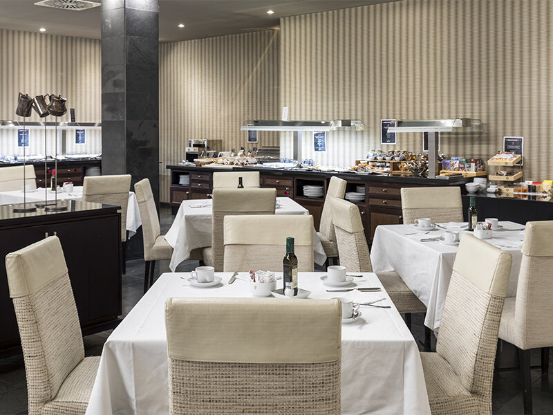 Un restaurante de hotel con mesas con manteles blancos, sillas beis y un bufé al fondo.