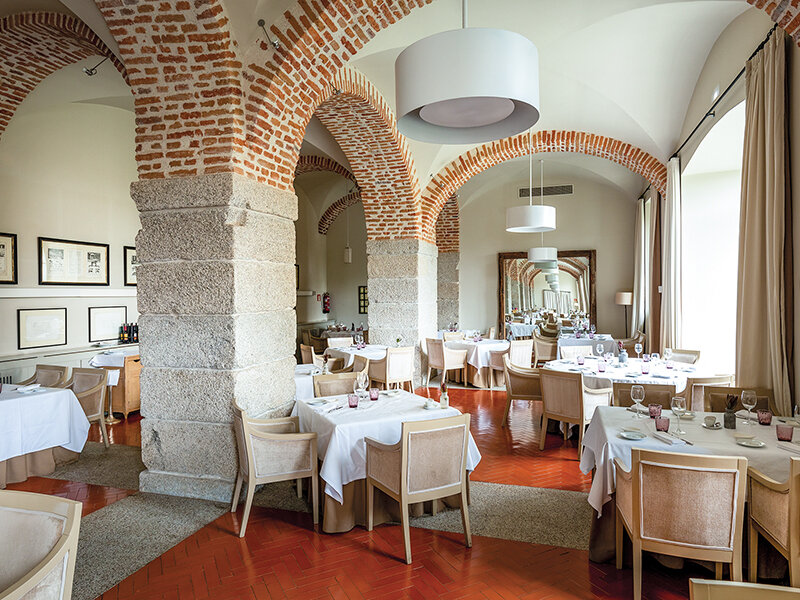 Un restaurante elegante con arcos de ladrillo rojo, pilares de piedra, suelos de madera y mesas con manteles blancos.