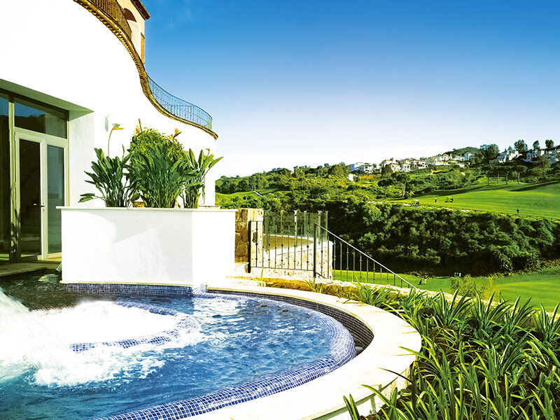 Un edificio blanco con piscina y jacuzzi domina un paisaje verde y montañoso.