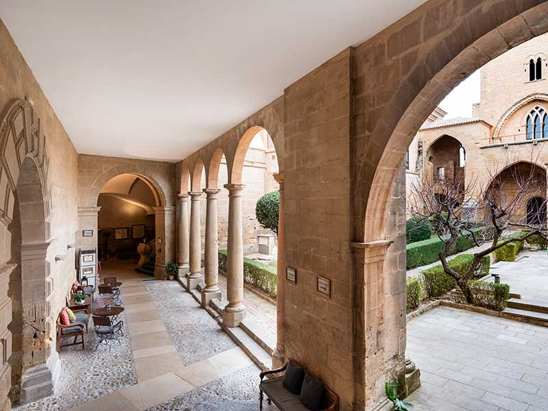 Un pasillo de arcadas de piedra con arcos que da a un patio con árboles y edificios históricos.