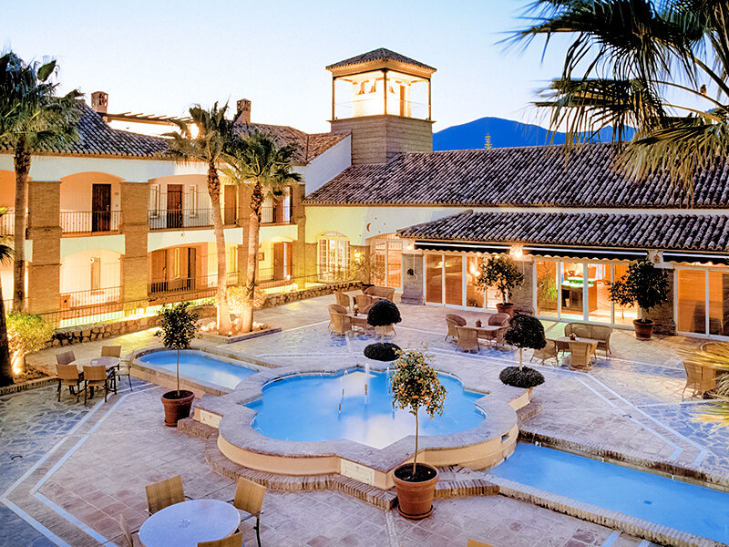 Un patio de hotel iluminado con una gran fuente, palmeras, mesas al aire libre y edificios de estilo mediterráneo.