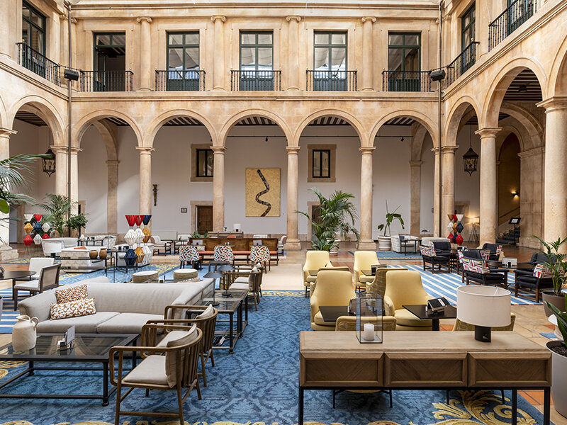 Espacioso patio interior con columnas beige, arcos, alfombras azules y muebles de salón modernos.