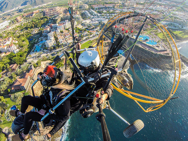 Vista aérea de dos personas volando en paramotor sobre una ciudad costera y el mar.