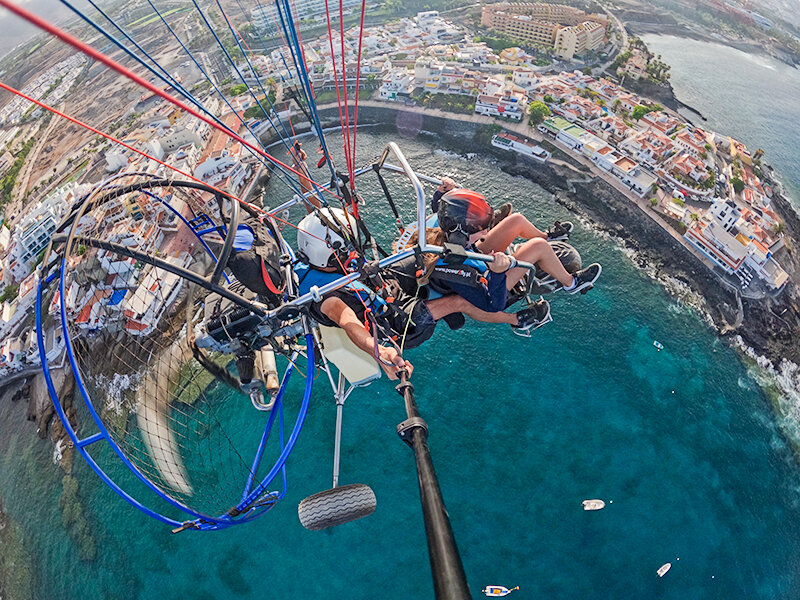 Dos personas vuelan en un paramotor sobre un pueblo costero con aguas cristalinas.