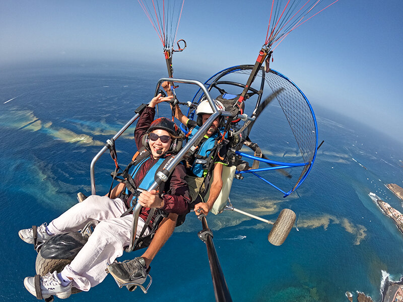 Dos personas vuelan en parapente a motor sobre el mar azul.