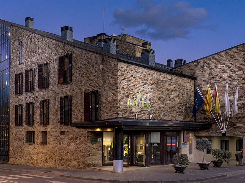 Un edificio de piedra rústica con letrero iluminado, fachada de cristal y banderas al atardecer.