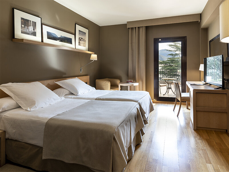 Una habitación de hotel moderna con dos camas, suelo de madera, un escritorio, televisión y un balcón con vistas.