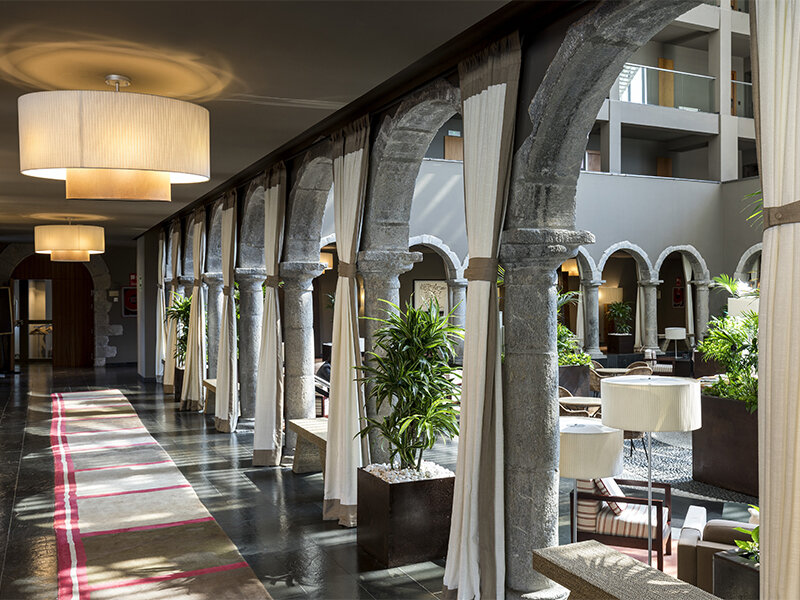 Un pasillo de hotel con arcos de piedra, cortinas blancas, lámparas colgantes y una alfombra estampada.