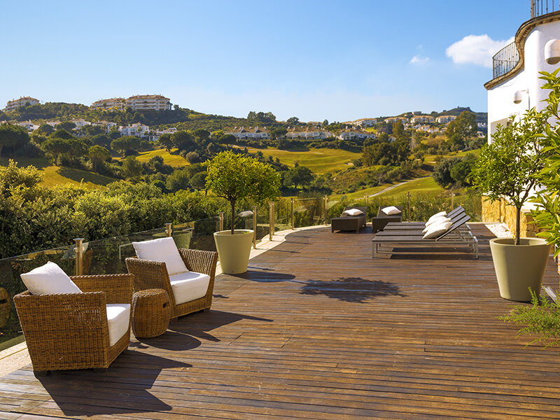 Soleada terraza de madera con sillones de mimbre, tumbonas y colinas verdes al fondo.