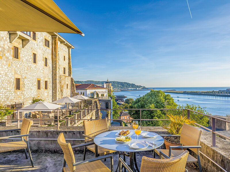Soleada terraza en la azotea con mesa, sillas de mimbre, desayuno y vistas al río, mar y edificios históricos.