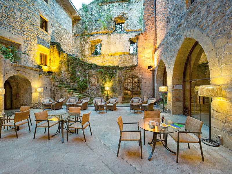 Un patio de piedra iluminado con mesas, sillas y lámparas rodeado de arquitectura histórica.