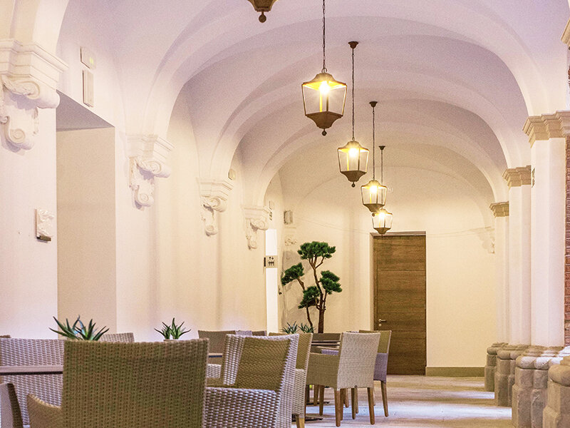 Un pasillo abovedado blanco con faroles colgantes, muebles de mimbre y una puerta de madera al fondo.