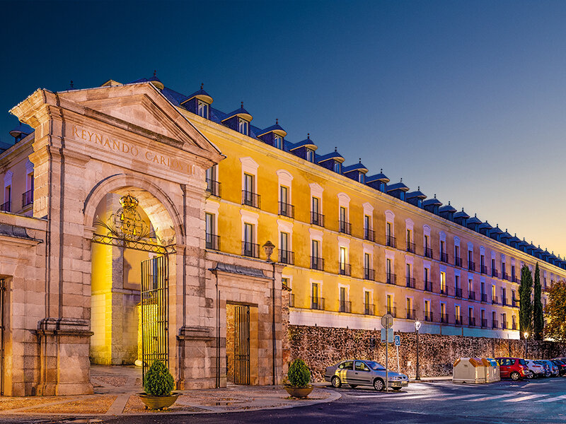 Puerta de piedra histórica iluminada ante un edificio amarillo largo al atardecer, con coches aparcados.