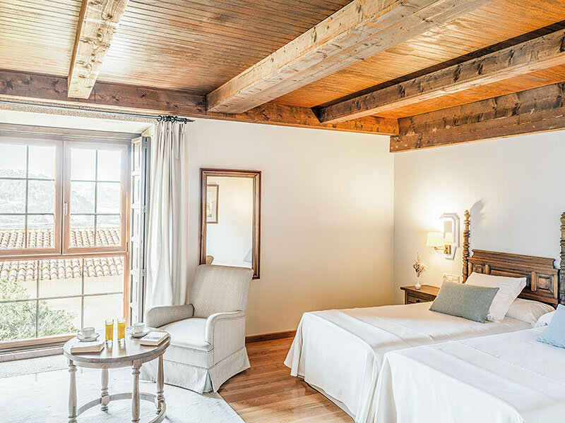 Acogedor dormitorio con vigas de madera, dos camas, sillón y una mesita con bebidas junto a la ventana.