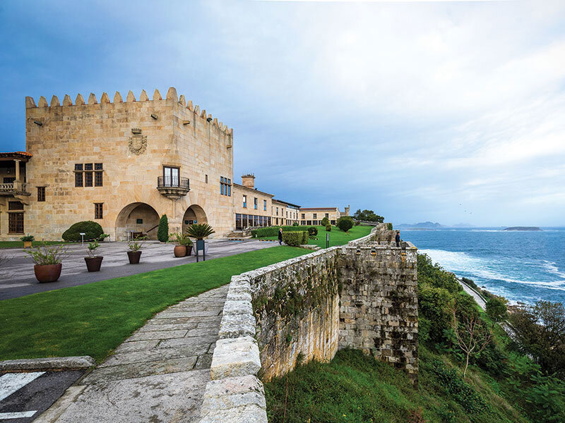 Un castillo de piedra histórico se encuentra junto a un césped verde y un muro de piedra junto al mar azul.