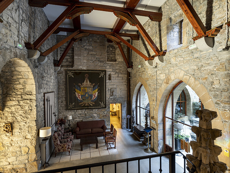 Salón de piedra con vigas de madera, tapiz, zona de estar y arcos que dan a un patio.