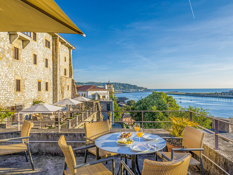 Terraza soleada con mesa puesta, vista al agua y edificio histórico de piedra bajo un cielo azul.