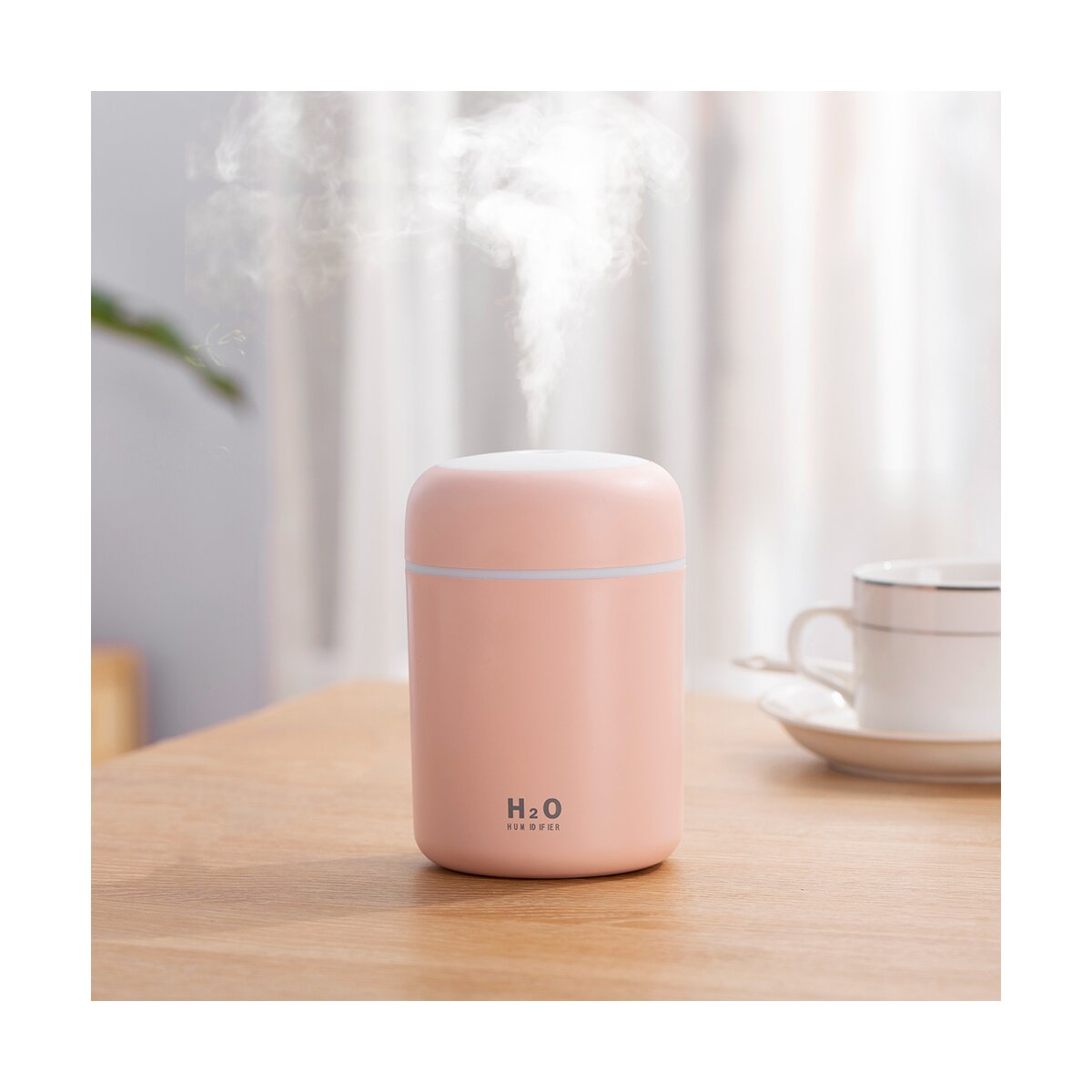 Ein rosa H2O-Luftbefeuchter auf einem Holztisch stößt weißen Nebel aus, mit einer weiße Tasse im verschwommenen Hintergrund.