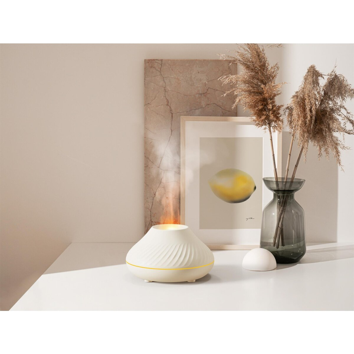 Ein weißer Diffusor mit sanftem Dampf steht auf einem Tisch, daneben eine Vase mit getrocknetem Gras und ein Bild.