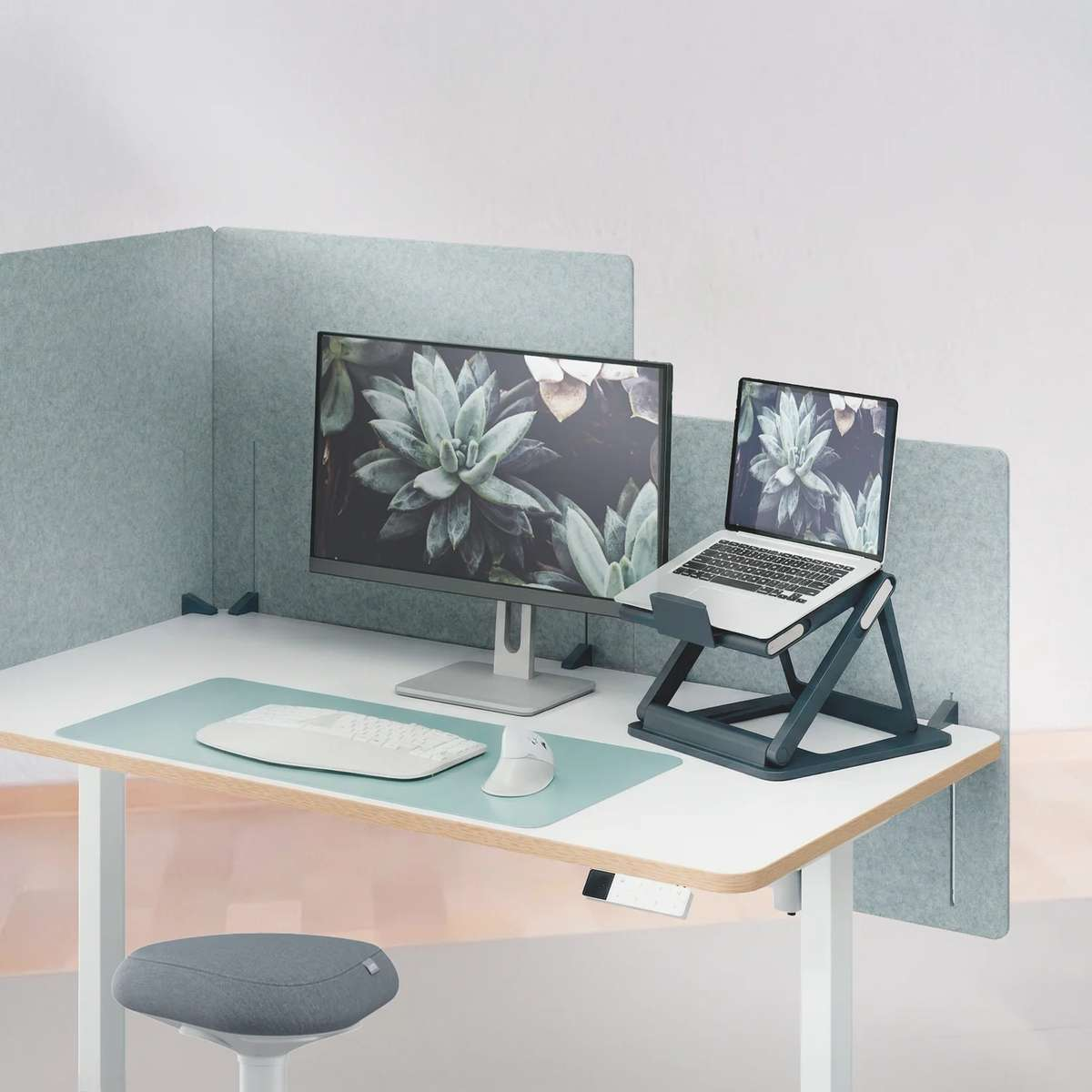Moderner Arbeitsplatz mit Laptop auf Ständer, Monitor, weißer Tastatur und Maus auf heller Matte mit Sichtschutz.