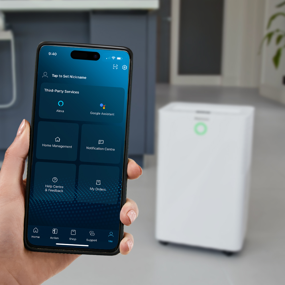Eine Hand hält ein Smartphone mit einer Smart-Home-App, im unscharfen Hintergrund ein weißer Luftentfeuchter.