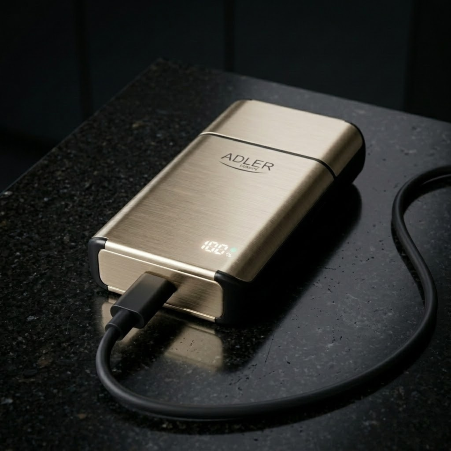 Złoty powerbank Adler z cyfrowym wyświetlaczem leży na ciemnym stole, podłączony jest do niego czarny kabel.