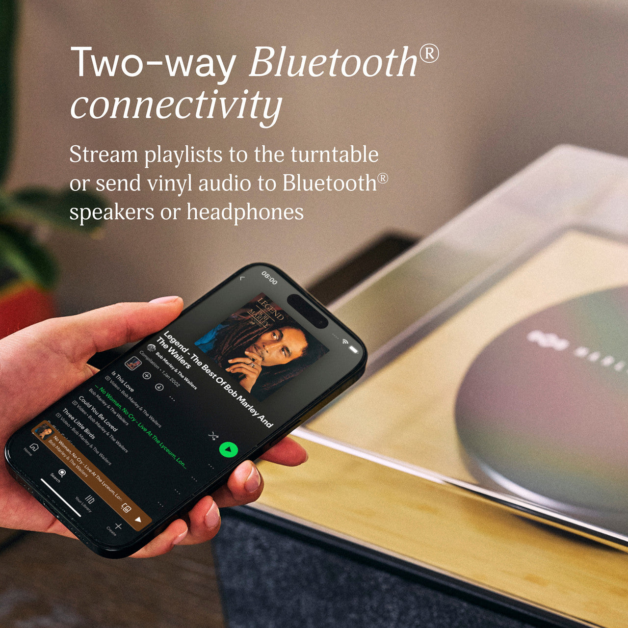 Hand hält Smartphone mit Musik-App, im Hintergrund ein Plattenspieler.