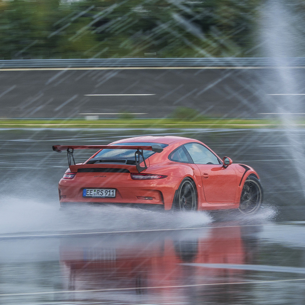 Ein roter Sportwagen driftet auf nasser Rennstrecke, Wasser spritzt und erzeugt Gischt.