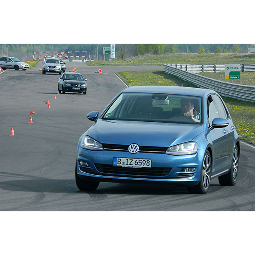 Ein blauer Volkswagen fährt auf einer Teststrecke mit weiteren Autos und Pylonen im Hintergrund.