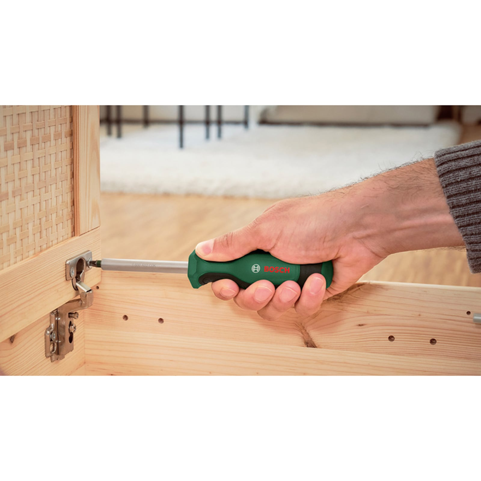 Eine Hand benutzt einen grün-schwarzen Bosch-Schraubendreher, um ein Scharnier an einem Holzkasten zu befestigen.