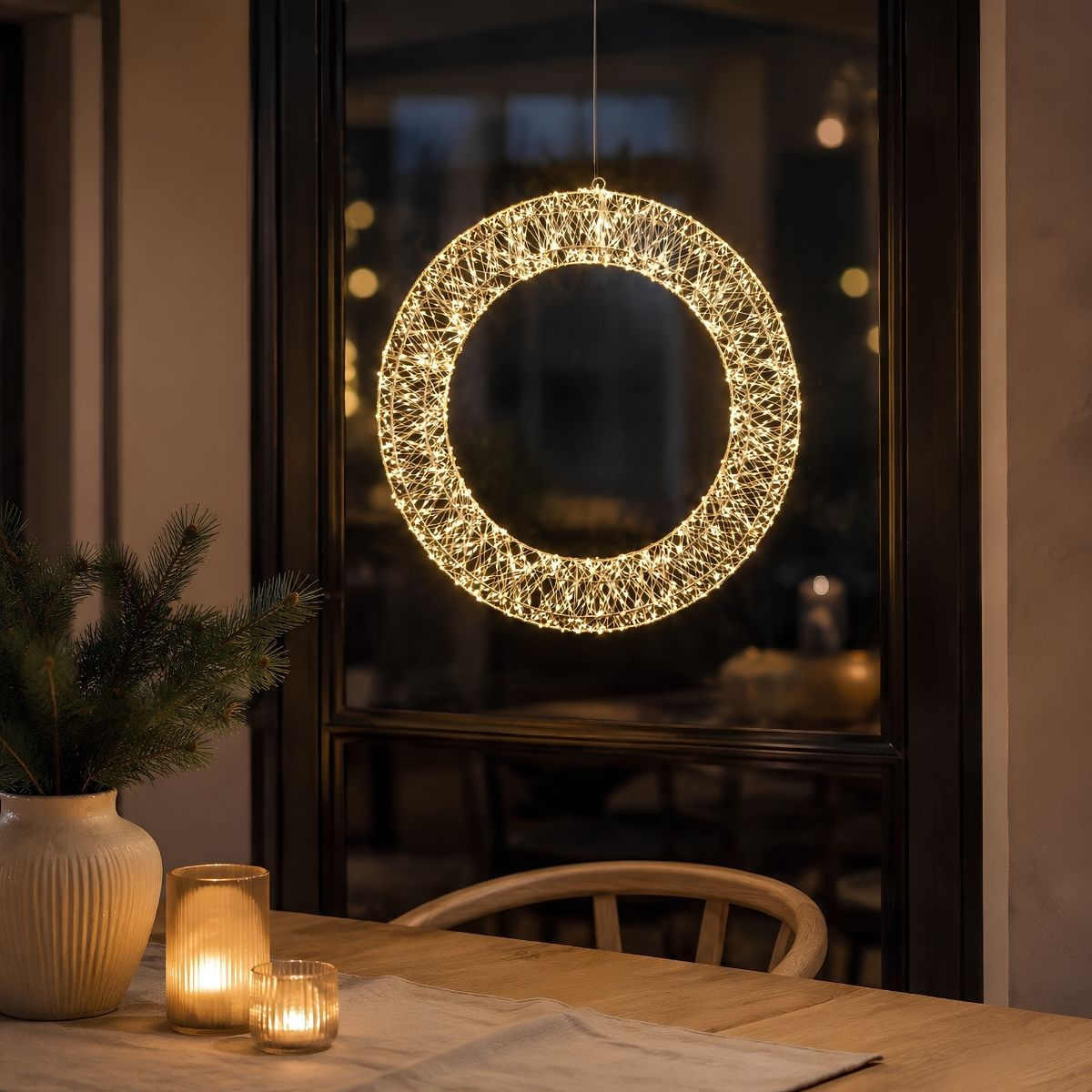 Een gloeiende lichtjeskrans hangt voor een raam boven een tafel met een vaas dennentakken en twee brandende kaarsen.