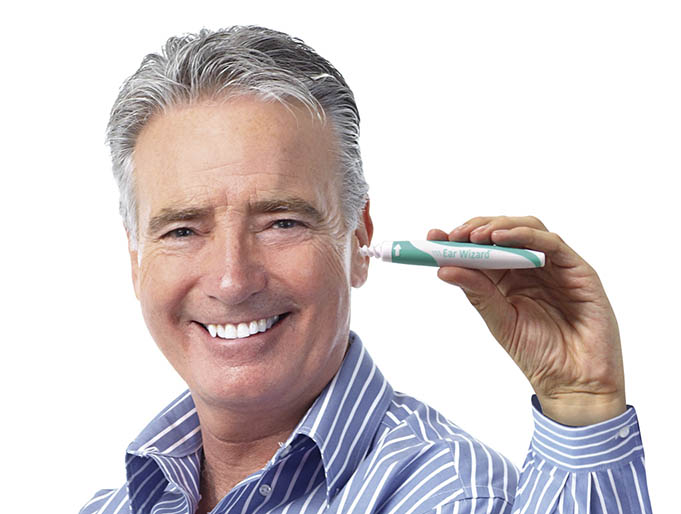 Ein älterer Mann in einem blau-weiß gestreiften Hemd lächelt und hält ein Ear-Wizard-Ohrpflegegerät an sein Ohr.
