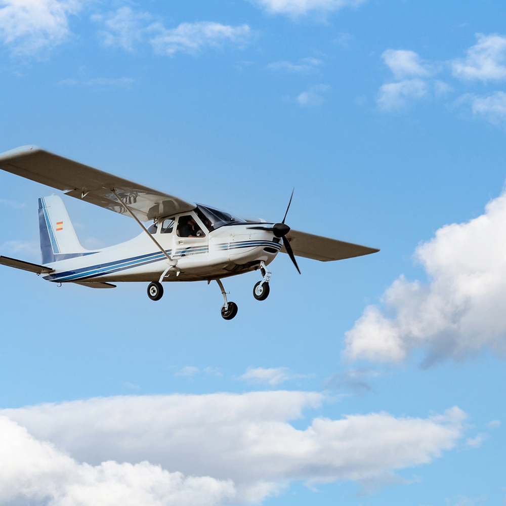 Ein kleines weißes Propellerflugzeug fliegt am blauen Himmel mit weißen Wolken.