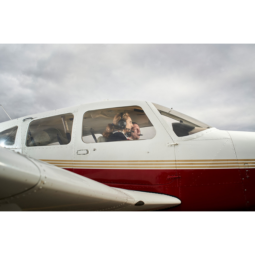 Ein Pilot und Co-Pilot mit Headsets sitzen im Cockpit eines kleinen weiß-roten Flugzeugs unter bewölktem Himmel.