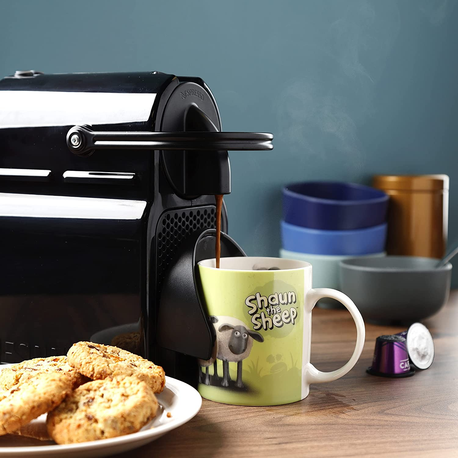 Una cafetera negra vierte café en una taza de Shaun el Cordero junto a galletas y cuencos apilados.