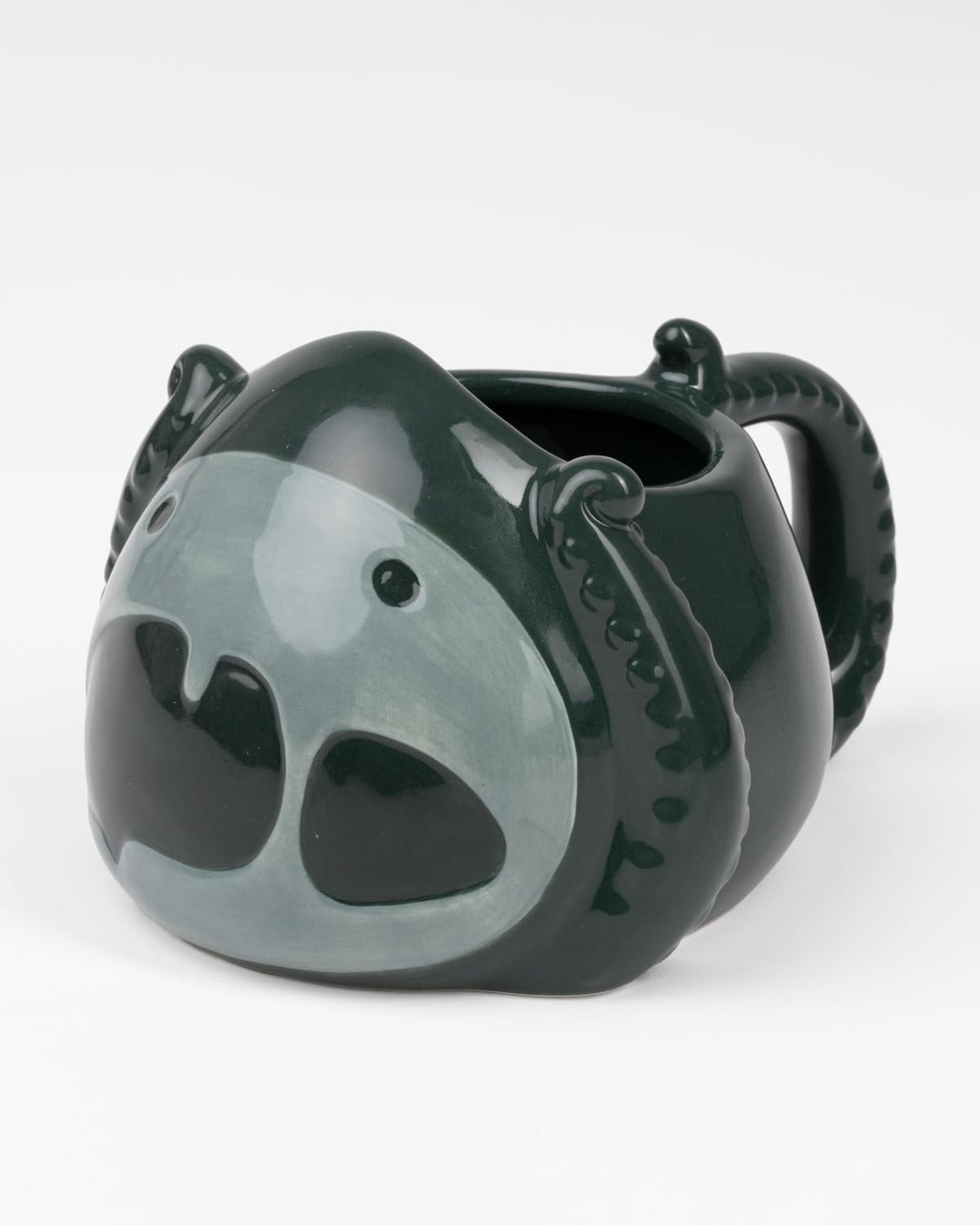 Taza de cerámica verde oscura con cara de animal, ojos grandes y asa de tentáculo, sobre fondo blanco.