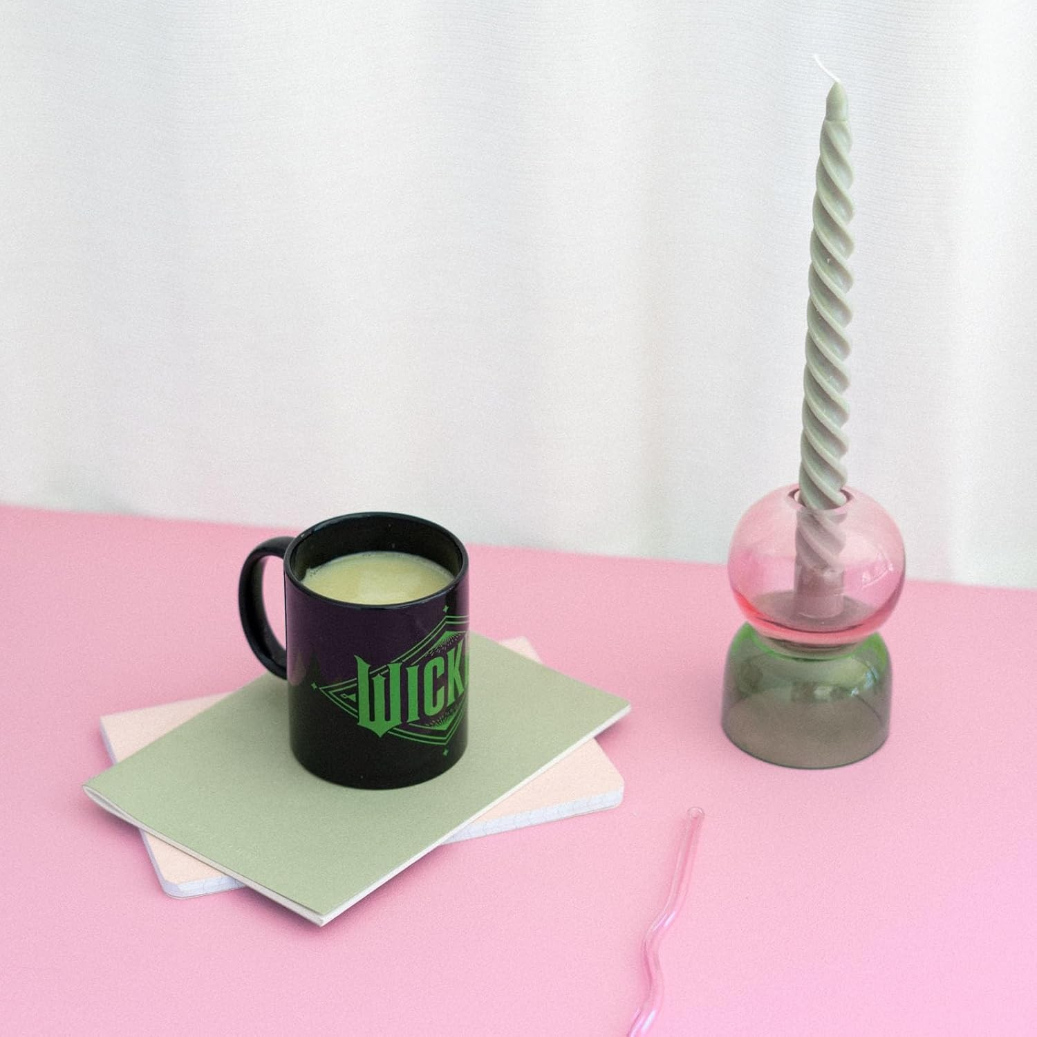 Una taza negra sobre cuadernos pastel junto a una vela torcida en un soporte de vidrio en una mesa rosa.