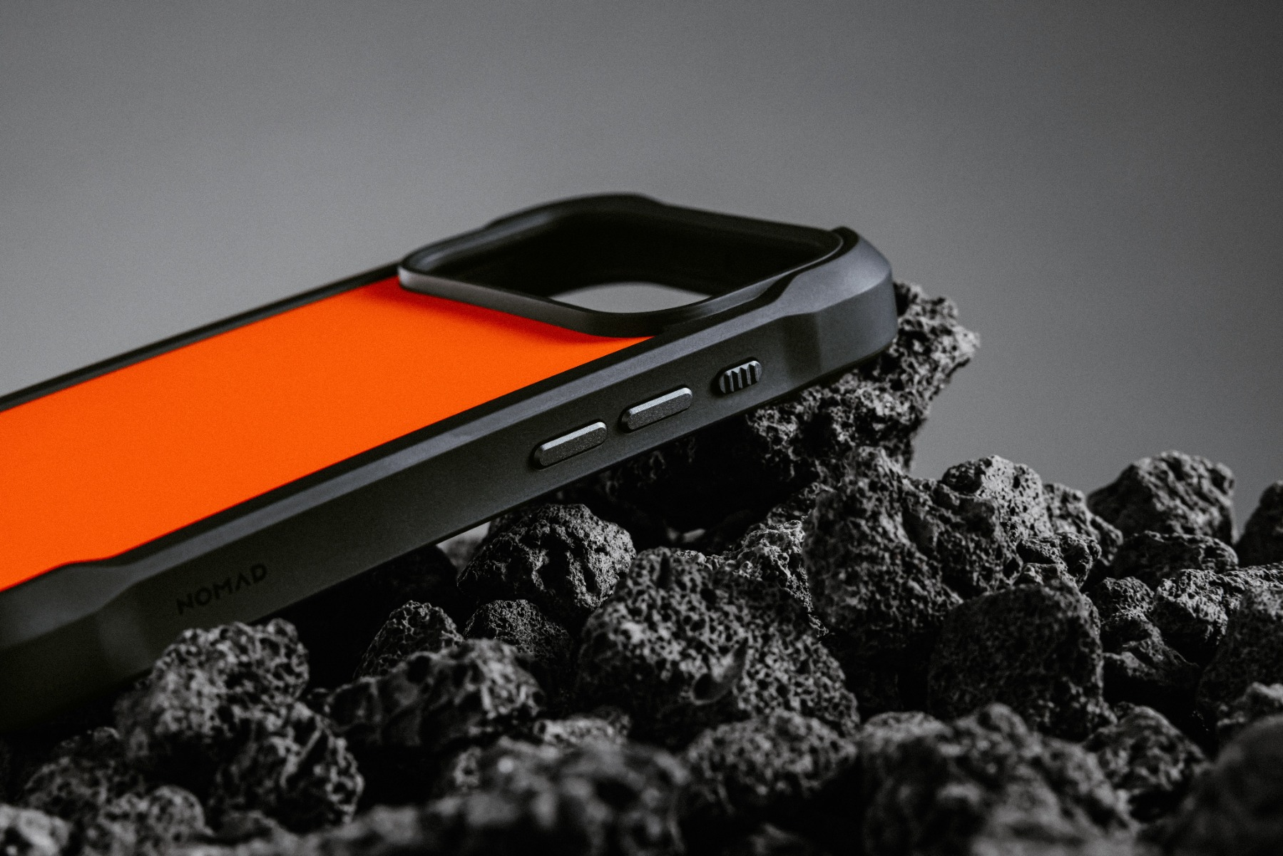 Czarno-pomarańczowe etui na smartfon Nomad spoczywa na ciemnych skałach lawowych na szarym tle.